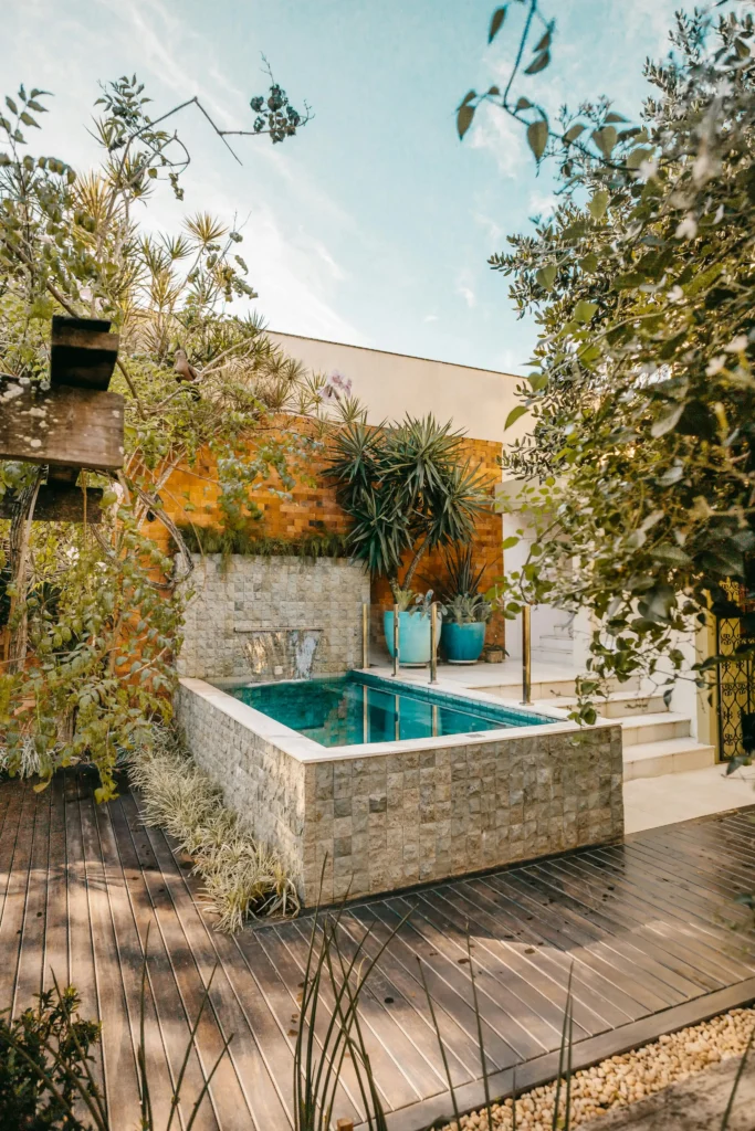 Elegant small plunge pool with stone-clad walls, cascading water feature, wooden deck flooring, and lush tropical landscaping in a modern outdoor courtyard.